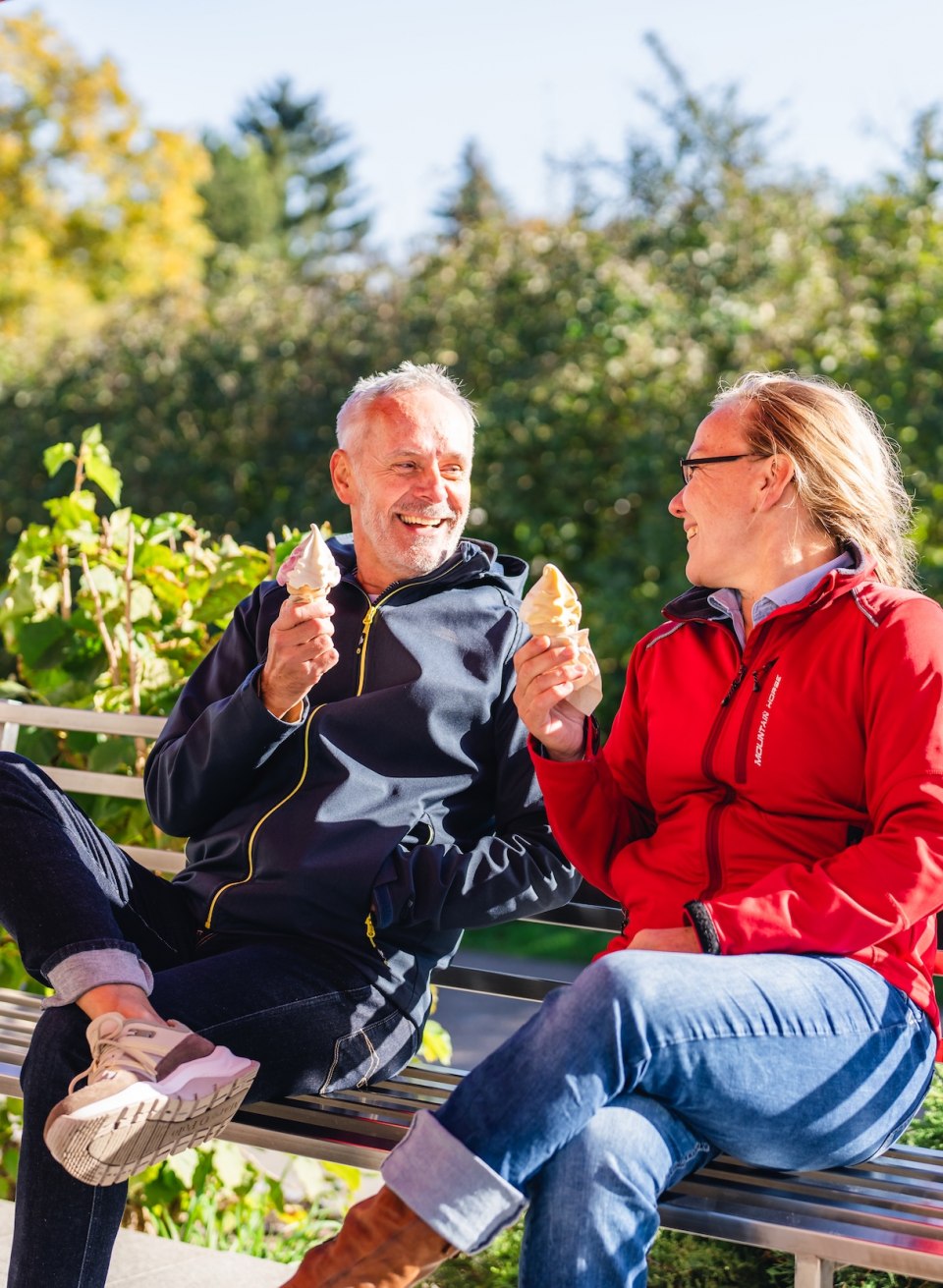 A couple enjoying soft ice cream on a bench in the sun, with green surroundings in the background.
