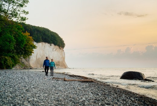 Een koppel wandelt langs de voet van de beroemde krijtrotsen op het eiland Rügen, terwijl de zonsondergang de hemel in zachte kleuren oplicht.