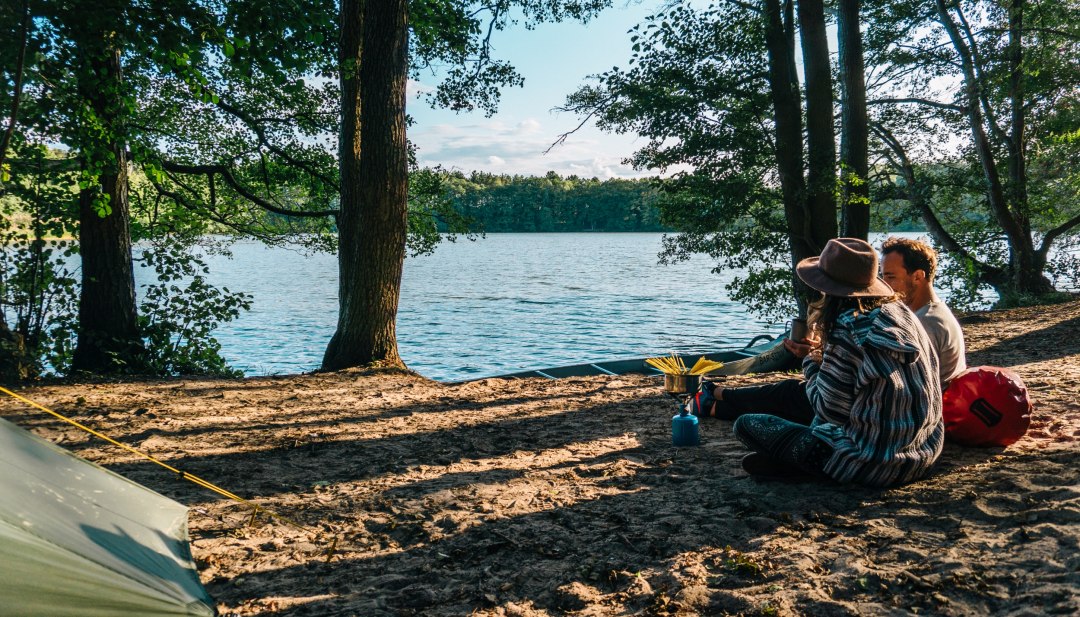 Een echtpaar zit aan het water met een gasfornuis en kookt pasta op camping Hexenwäldchen