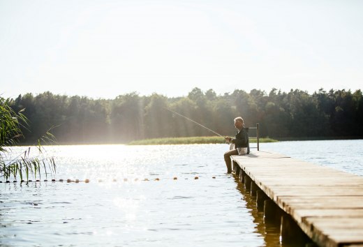 An elderly man sits on a wooden jetty by a calm lake and fishes while the sun makes the water glisten.