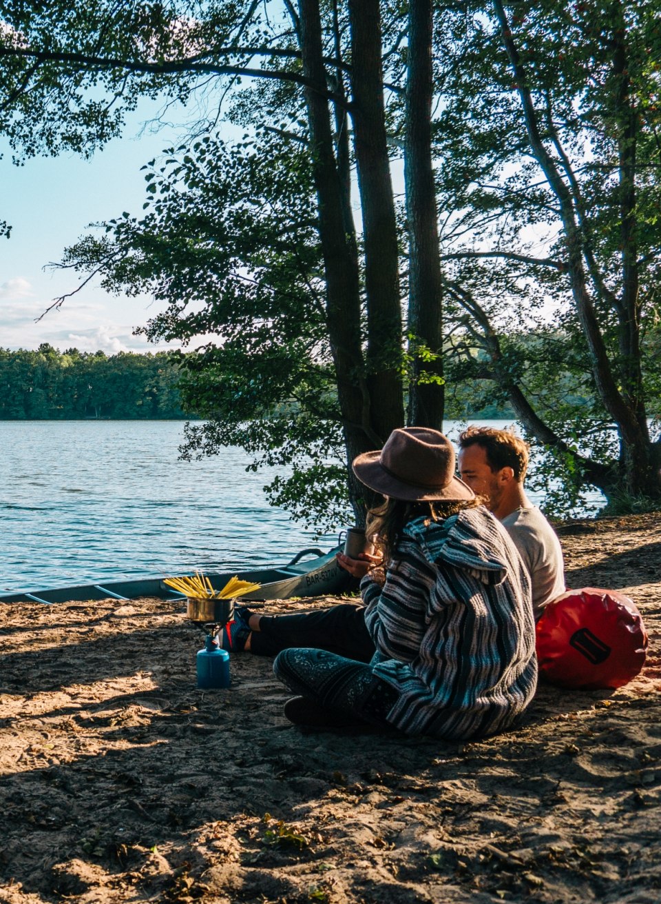 A couple sits by the water with a gas stove and cooks pasta at the Hexenwäldchen campsite