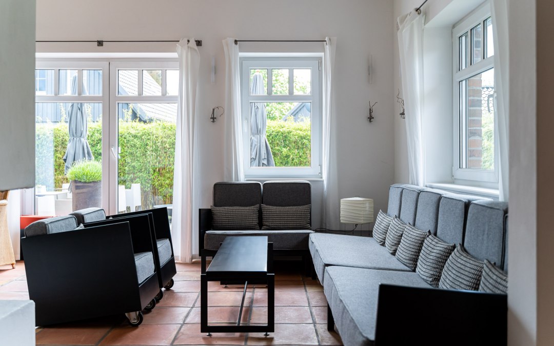 A bright and stylishly furnished lounge area with large windows that let in plenty of natural light. Gray sofas and armchairs with minimalist black accents invite you to relax. The warm terracotta floors and simple white curtains give the room a relaxed and inviting atmosphere., © Hotel Carpe Diem