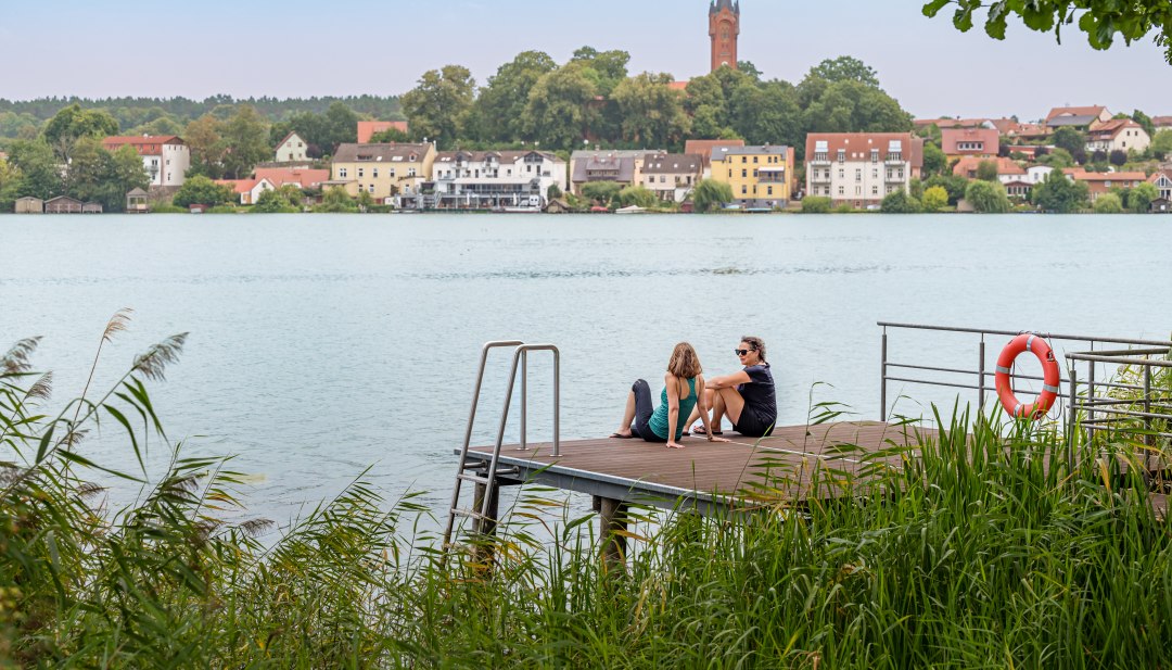 Two women sit on a jetty and look out over a lake