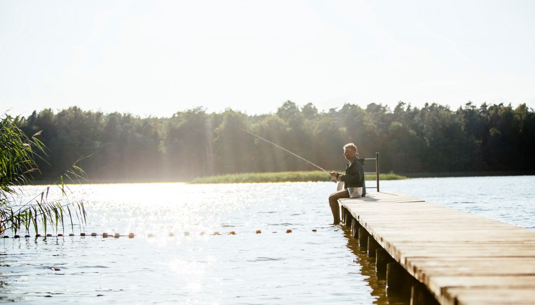 An elderly man sits on a wooden jetty by a calm lake and fishes while the sun makes the water glisten.
