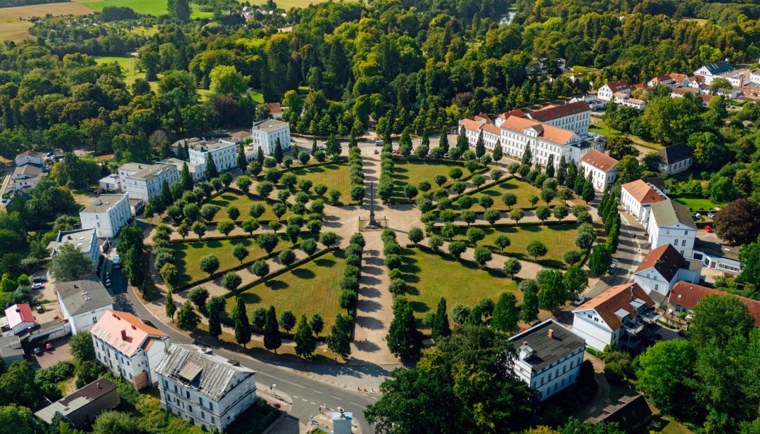 Aerial view of the Circus in Putbus on the island of Rügen, a circular square with symmetrically arranged trees and classicist buildings, surrounded by green nature and a rural landscape.