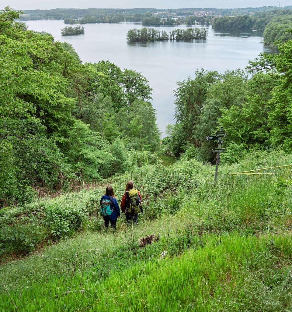 Two women hike along the nature park trail and look down from the mountain onto the Schmaler Luzin lake.